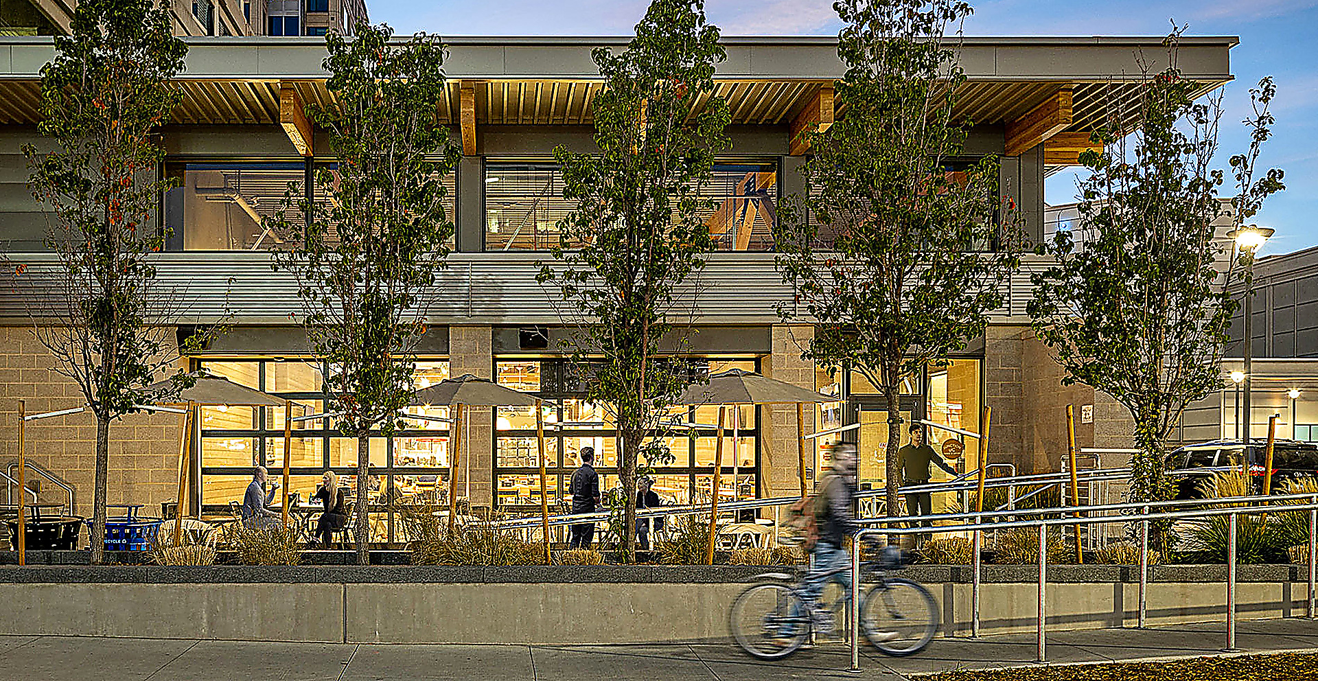 Junction Food Hall at dusk — a light masonry building with roll-up glass doors glowing warm from inside, umbrellas and patrons on the patio, slender trees planted along the sidewalk, a twilight sky overhead