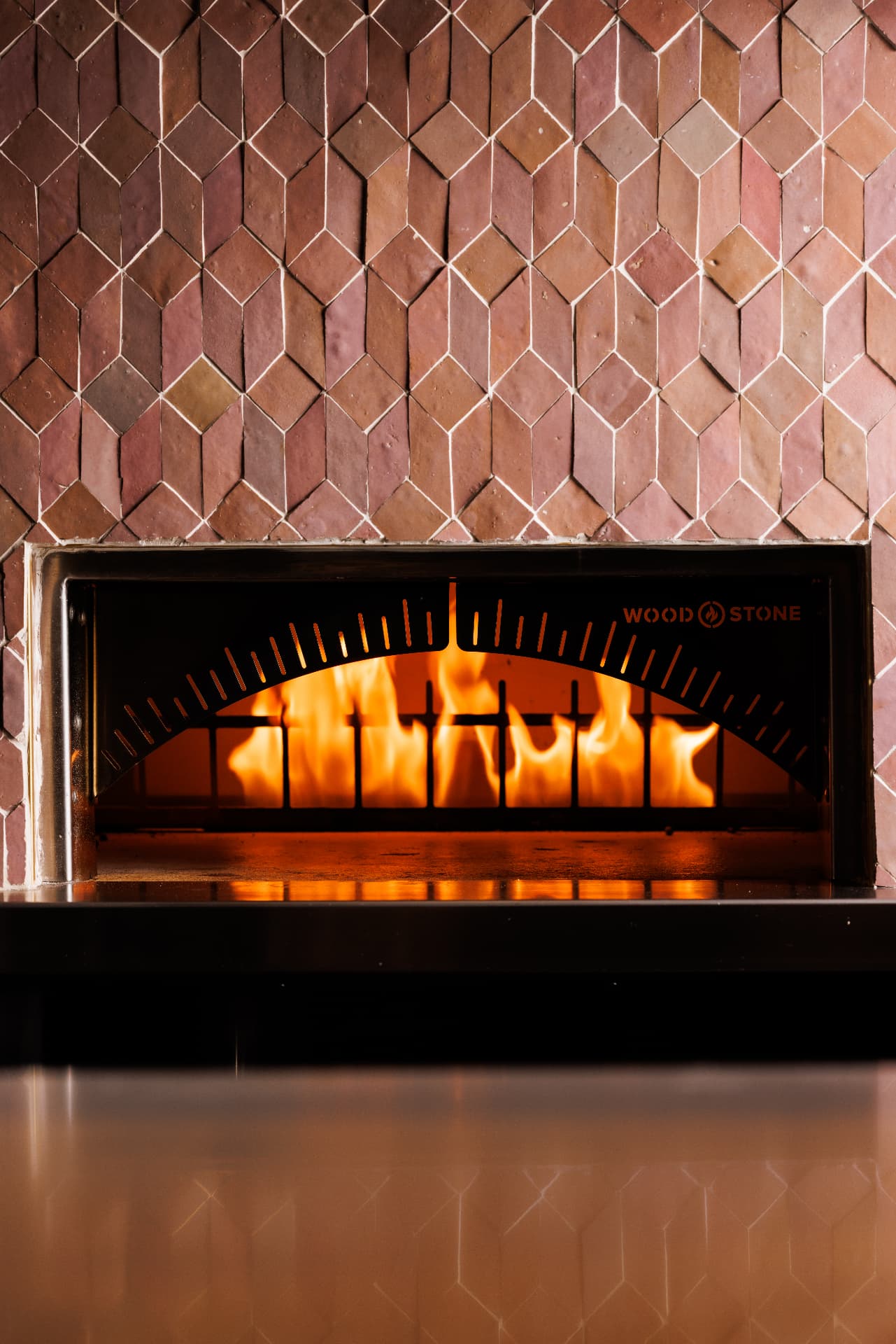 Wood-fired oven blazing behind a black steel arch with glowing temperature markings, set into a diamond-patterned tile wall of rose, terracotta and cream above a polished steel counter