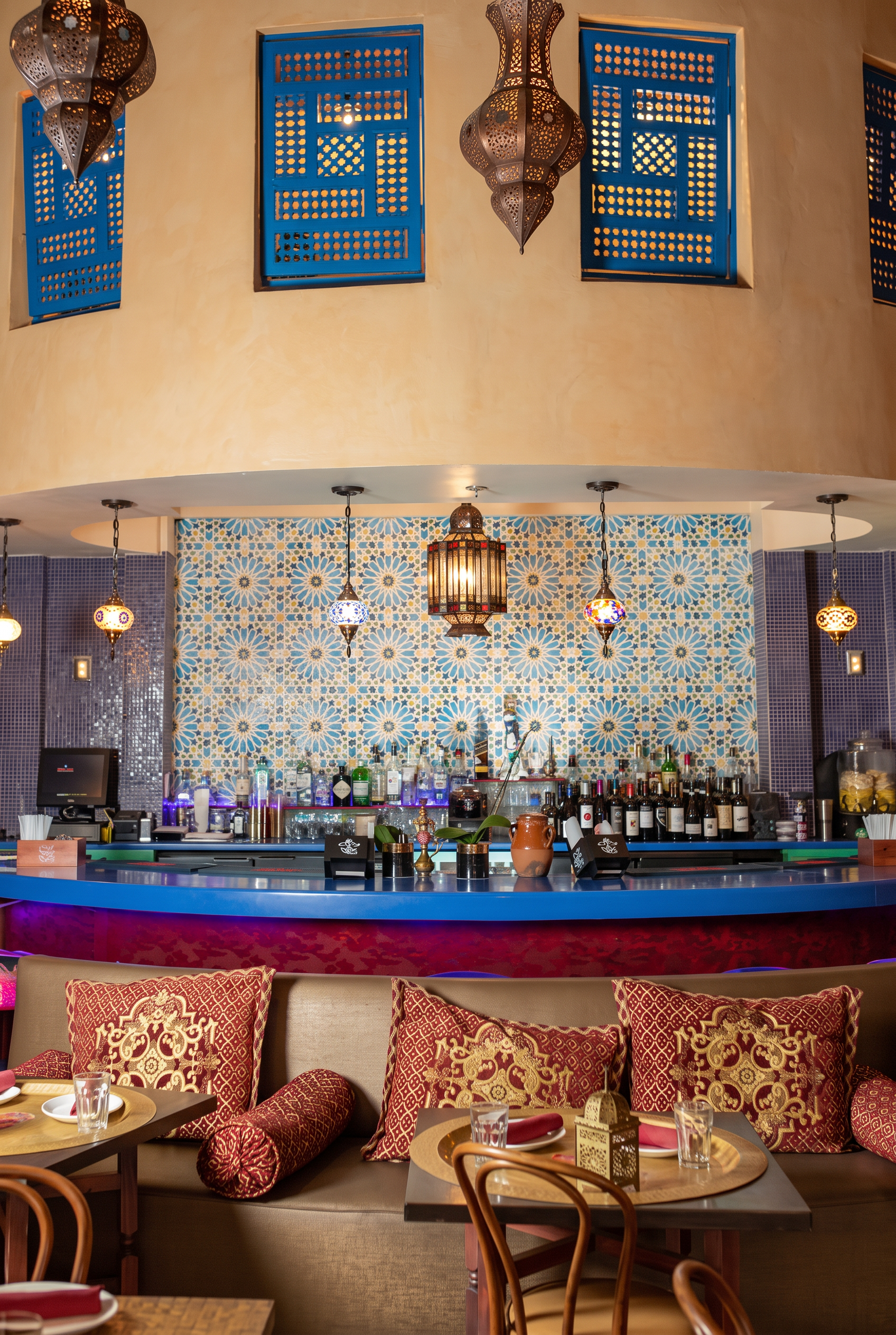The bar at night — blue mosaic tile, magenta glow beneath the counter, pierced metal lanterns, and a banquette dressed in gold and red embroidered pillows