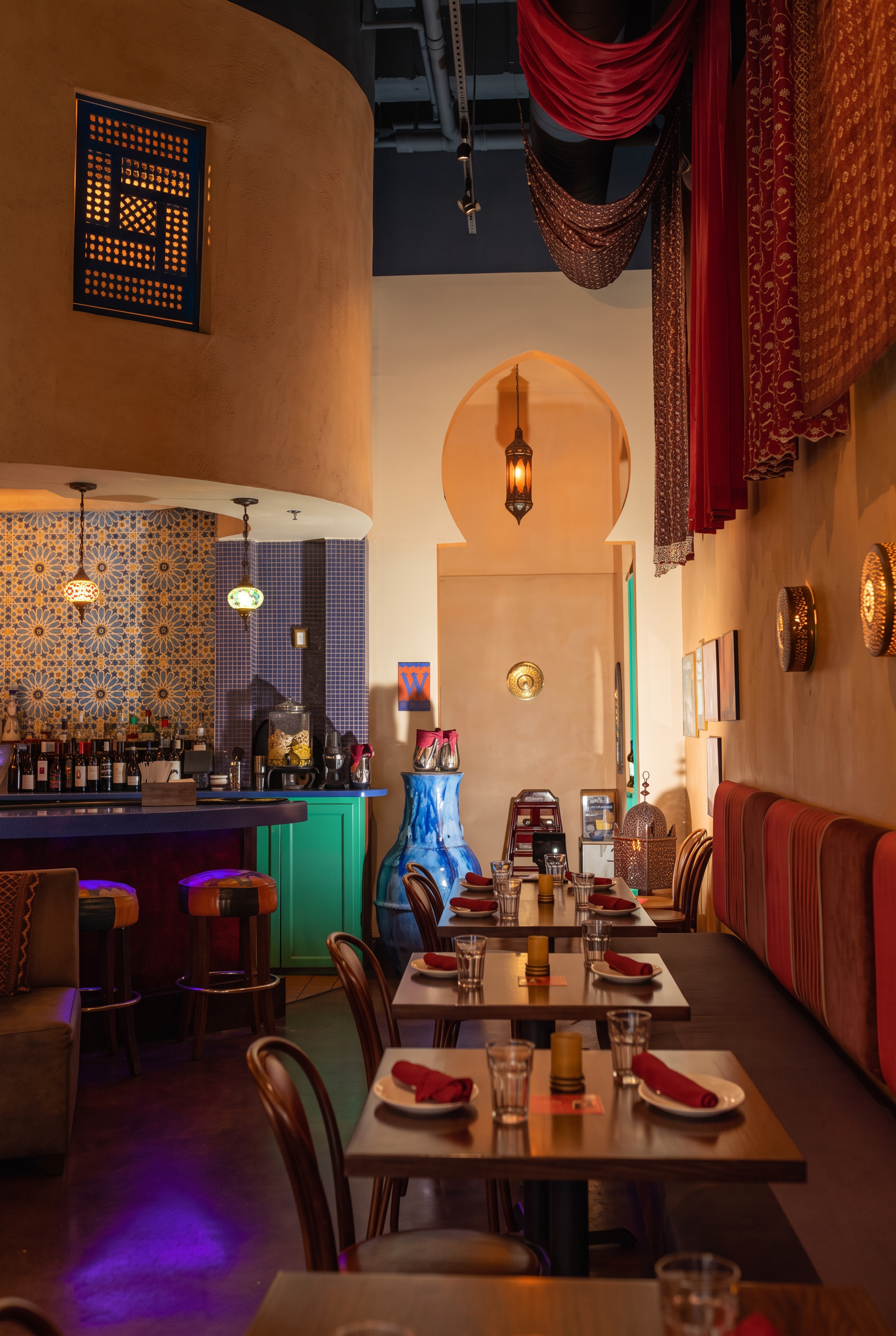 Evening dining room with terracotta plaster walls, a ribbed rust-coloured banquette and bentwood chairs, a mosaic-tiled bar to the left, draped crimson canopies, and a Moorish archway lit by pierced brass lanterns