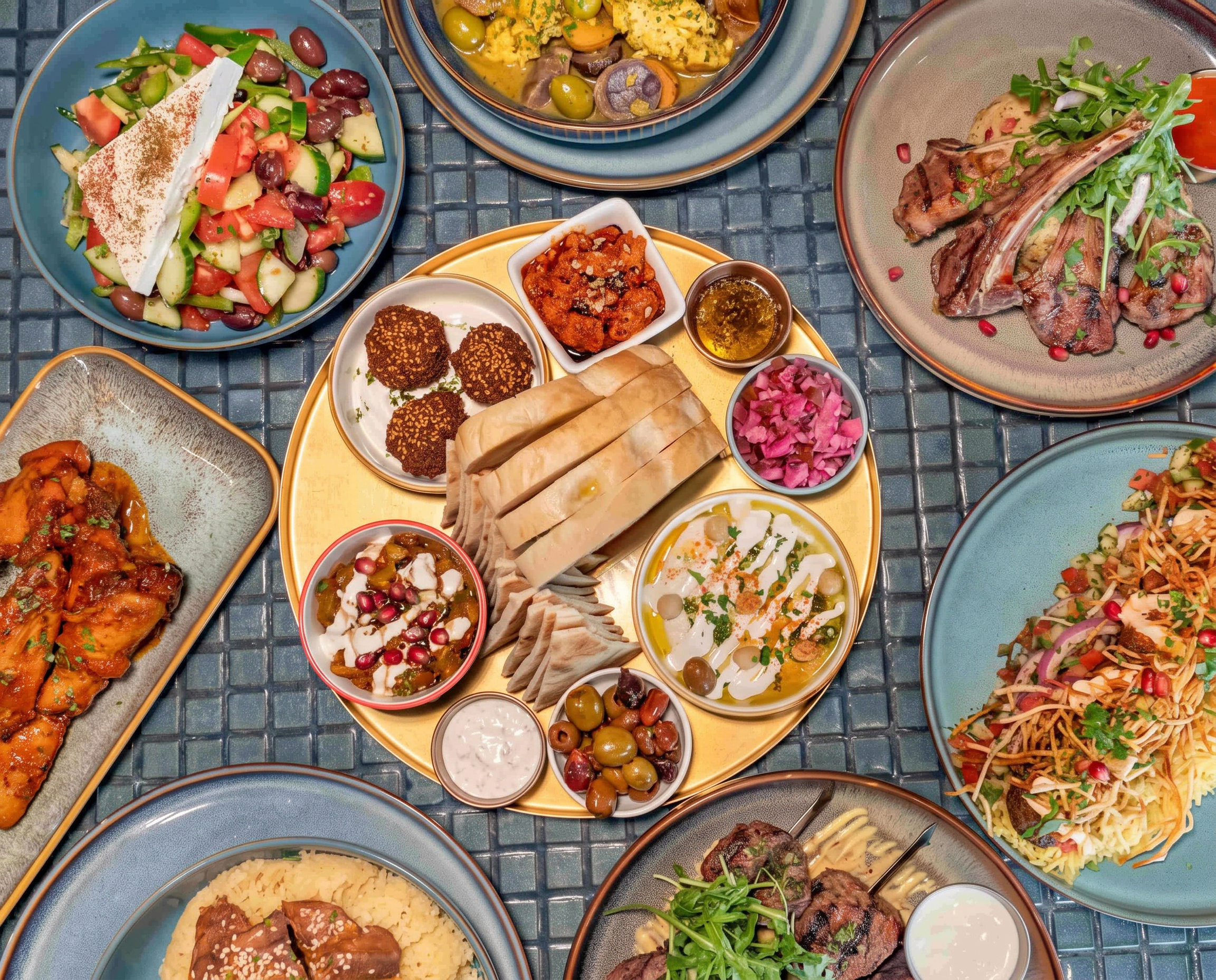 Overhead view of a mezze spread on a teal mosaic-tiled table — a brass platter of pita, dips and falafel, lamb chops, tagine, grilled skewers and rice dishes