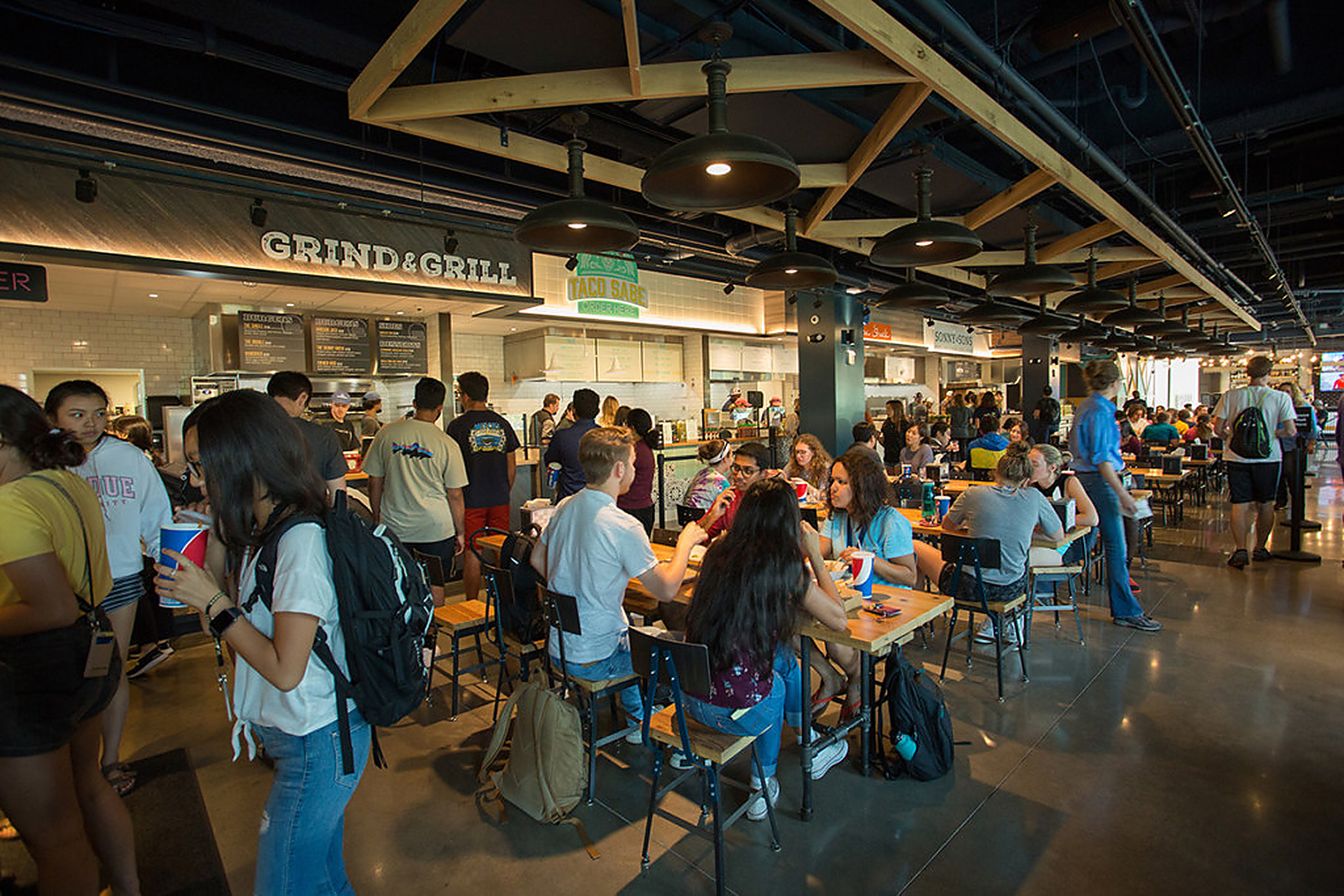 Students at long tables in front of the Grind & Grill stall, beneath timber trusses and rows of black dome pendants