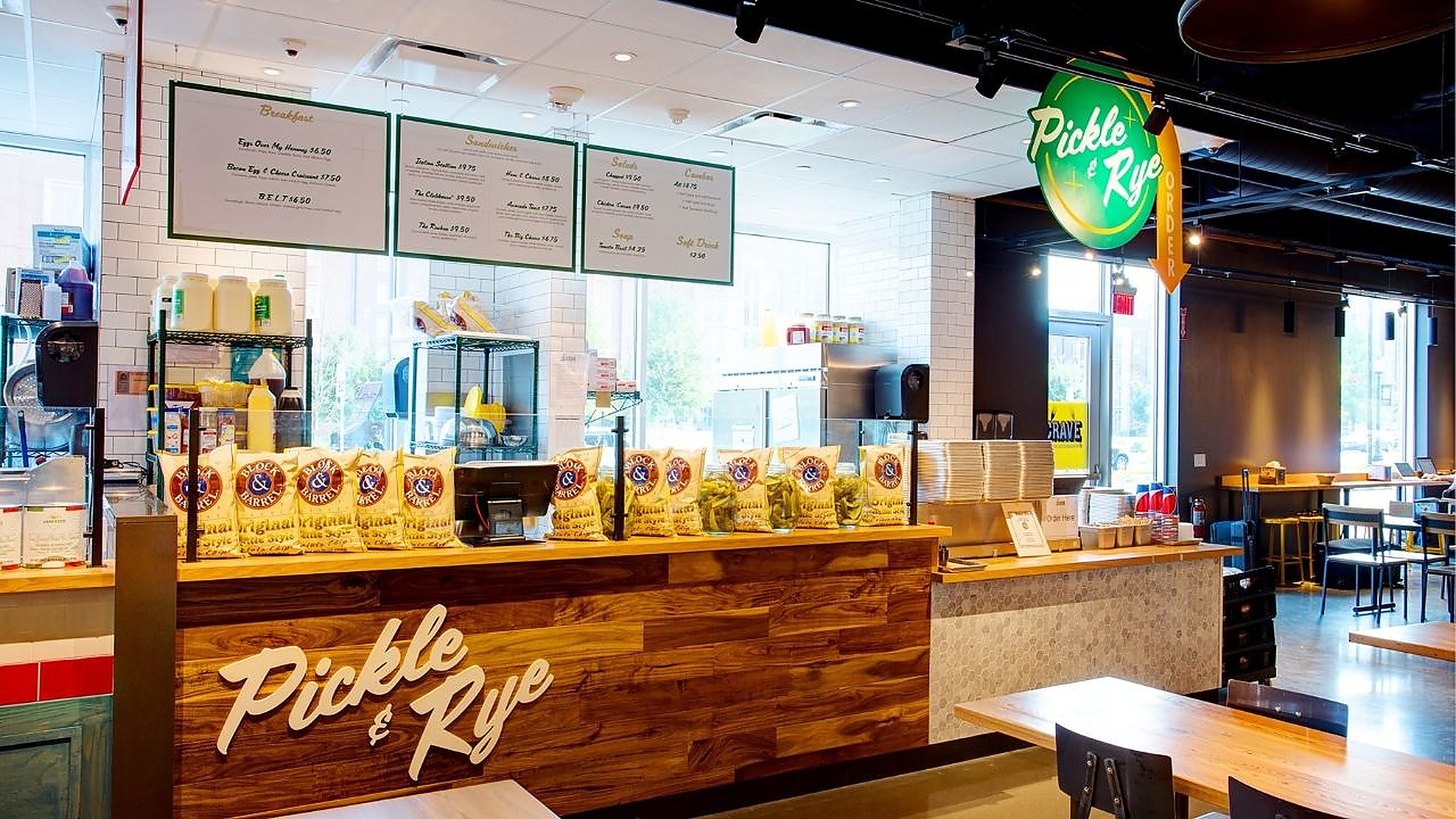 The Pickle & Rye deli counter at Crave — a wood-planked front with jars of dill pickles and bags of chips on the butcher-block top, green menu boards overhead, and the seating area beyond
