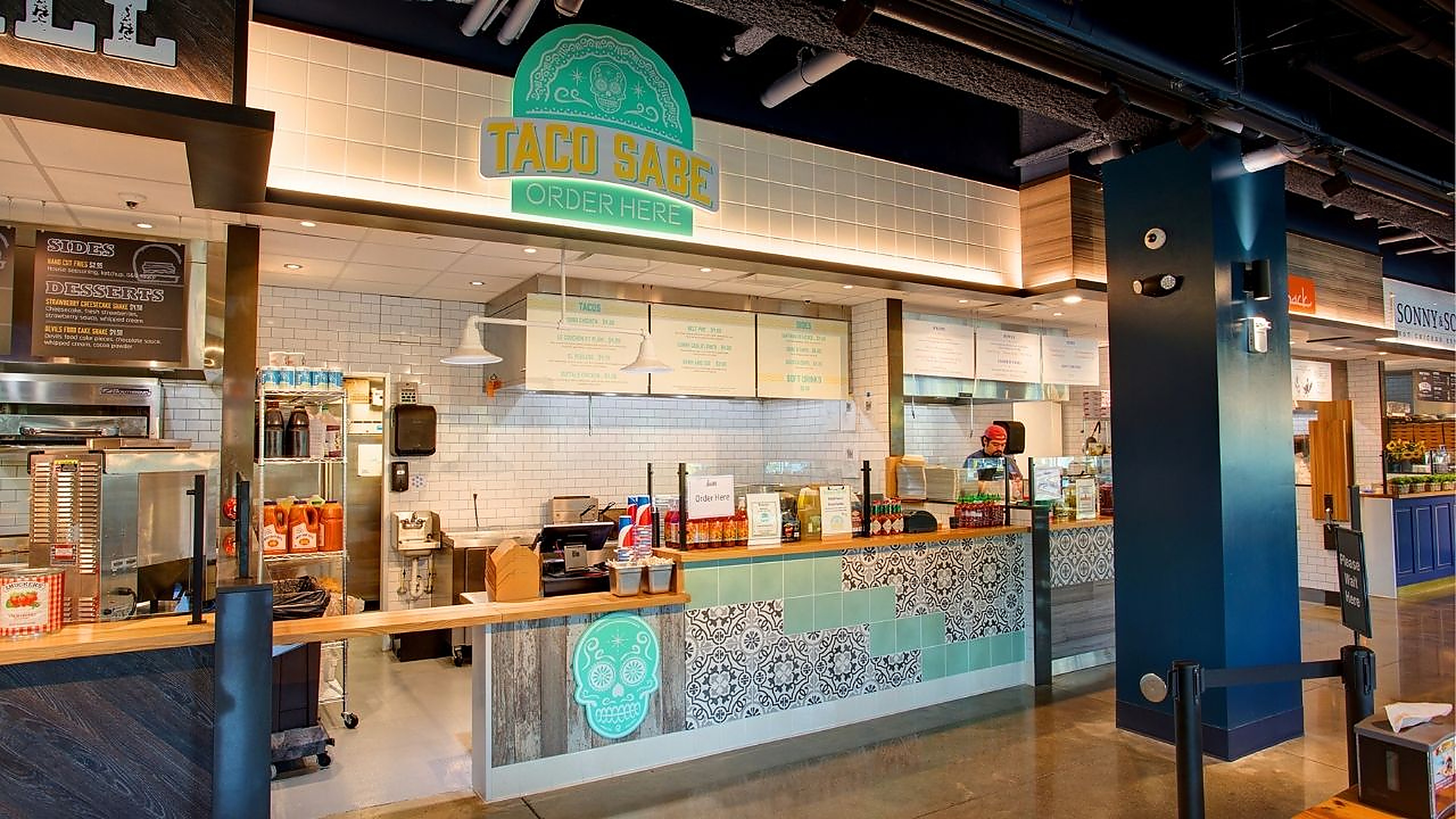 The Taco Sabe stall at Crave — a mint-tiled counter with a patterned-tile base, queue stanchions and an illuminated order marquee, opening onto a stainless-steel kitchen