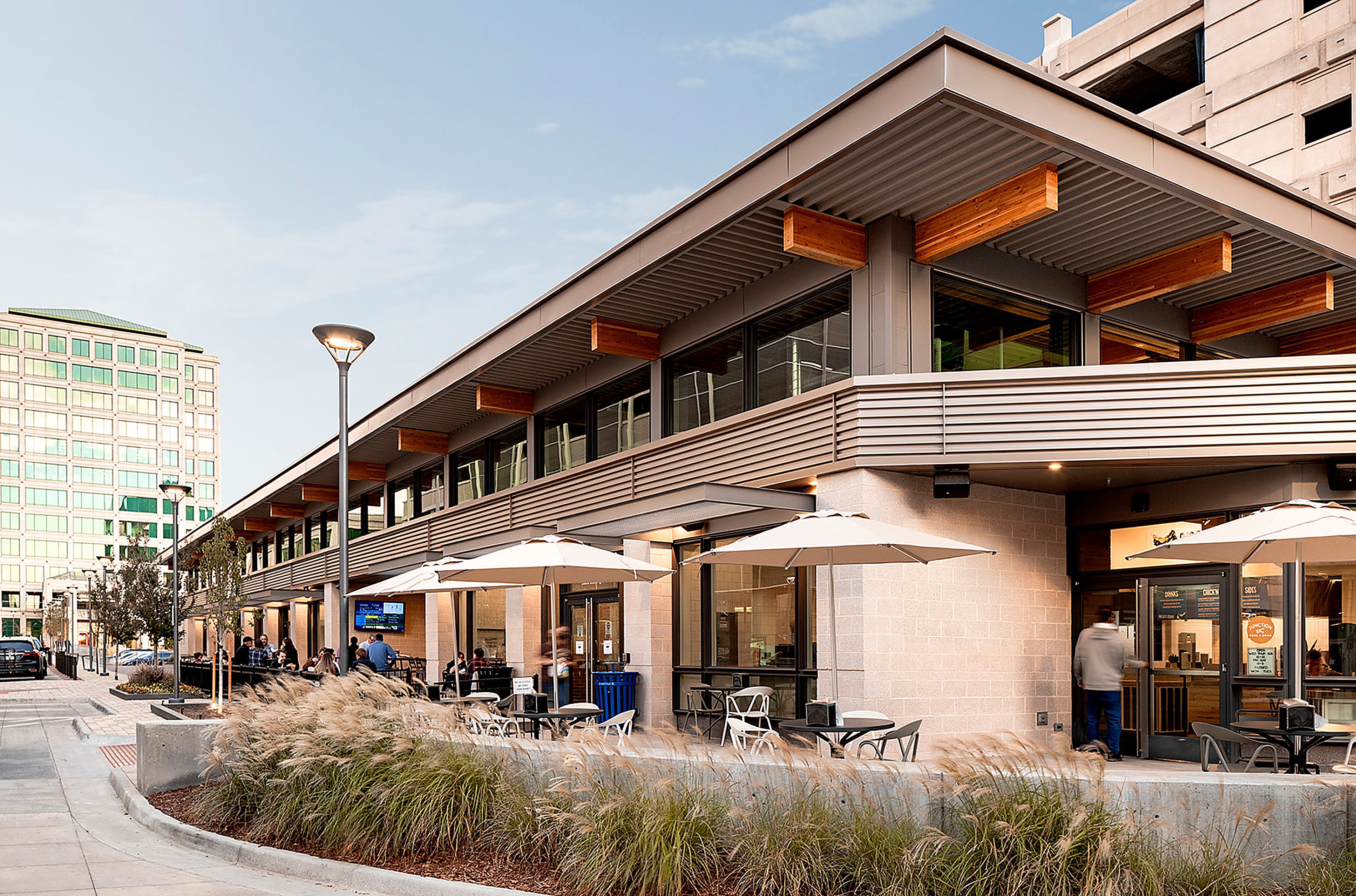 The corner of Junction at street level — café tables and beige umbrellas on the patio beside a curved concrete planter filled with tall ornamental grasses