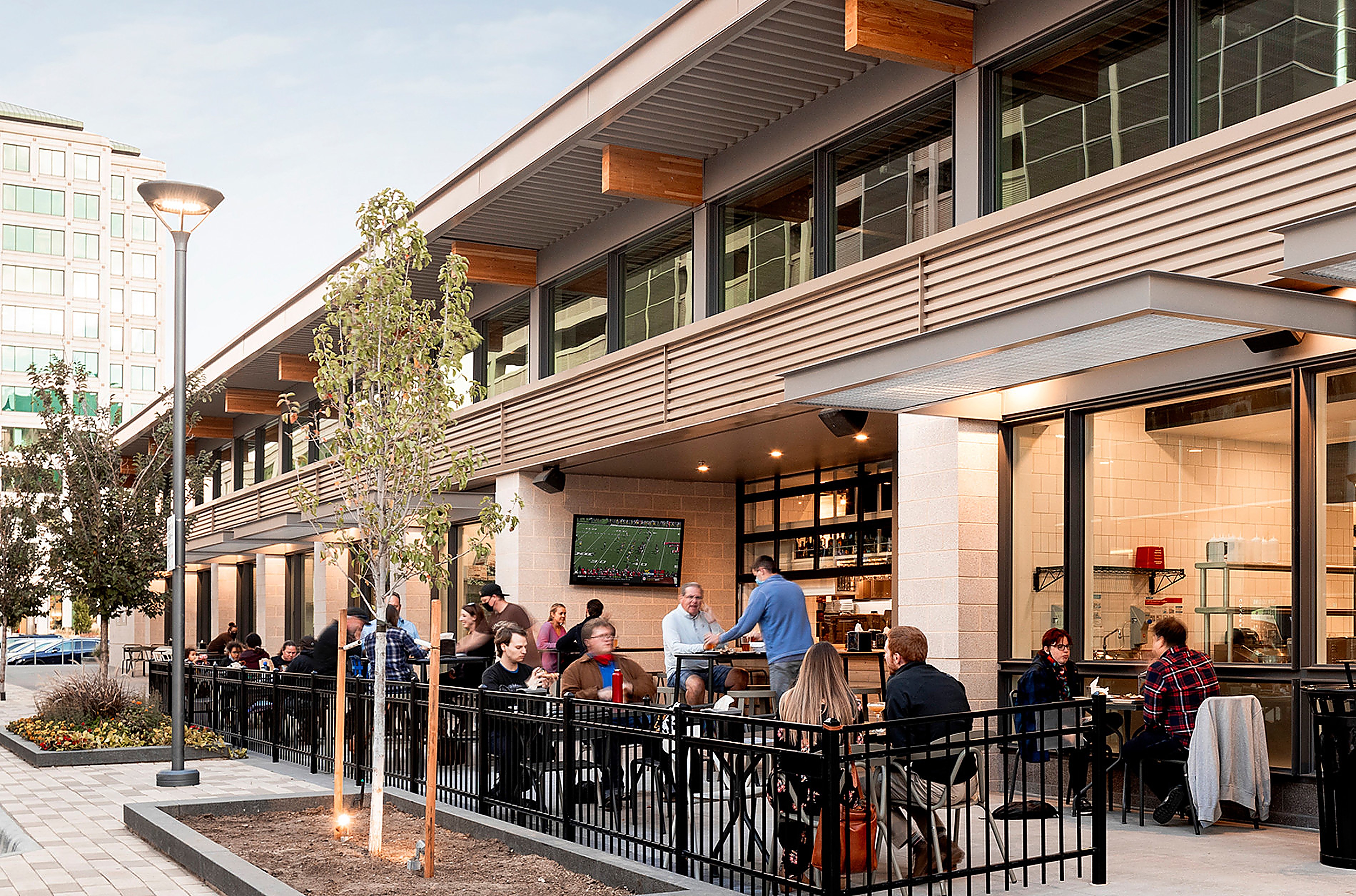 The side patio at Junction — patrons at timber tables beneath a wall-mounted television showing a game, with outdoor heaters and a black steel fence running along the ribbed metal wall