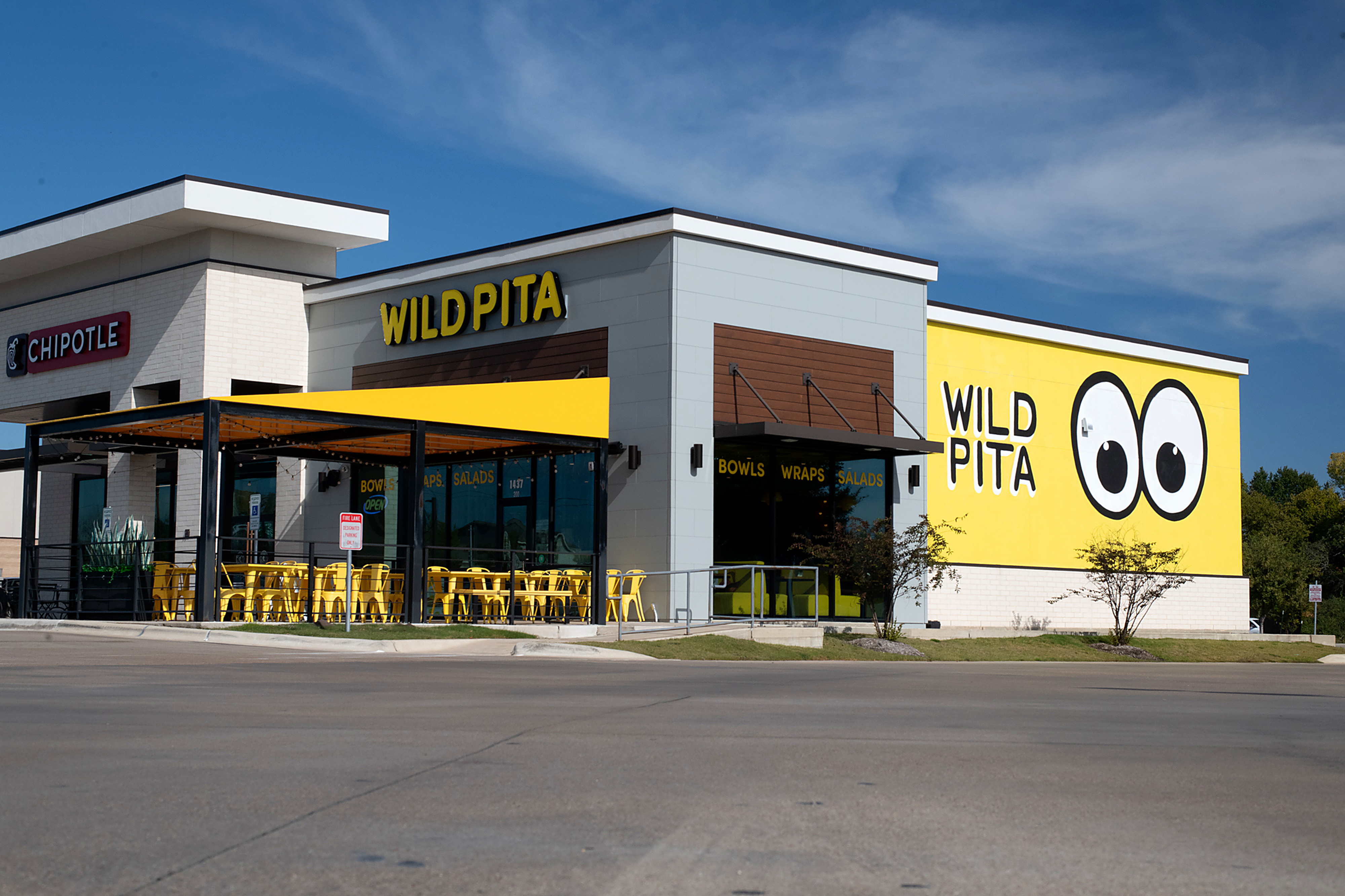 Wild Pita storefront at midday — a grey panel and wood-slat facade beside a bright yellow wall carrying the brand name and a googly-eye graphic, with a black steel patio canopy and yellow chairs out front