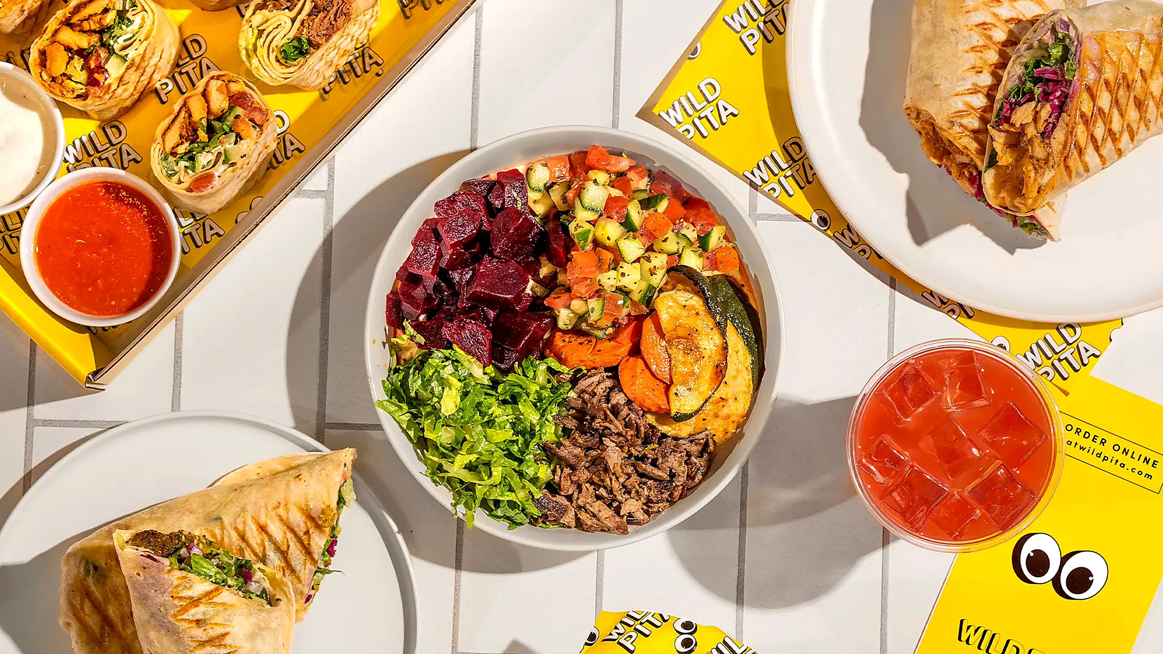 Overhead view of a Wild Pita spread — a grain bowl with beets, cucumber salad, grilled zucchini and shredded meat, sliced grilled pita wraps, small dips, and an iced red drink on bright yellow branded wrappers