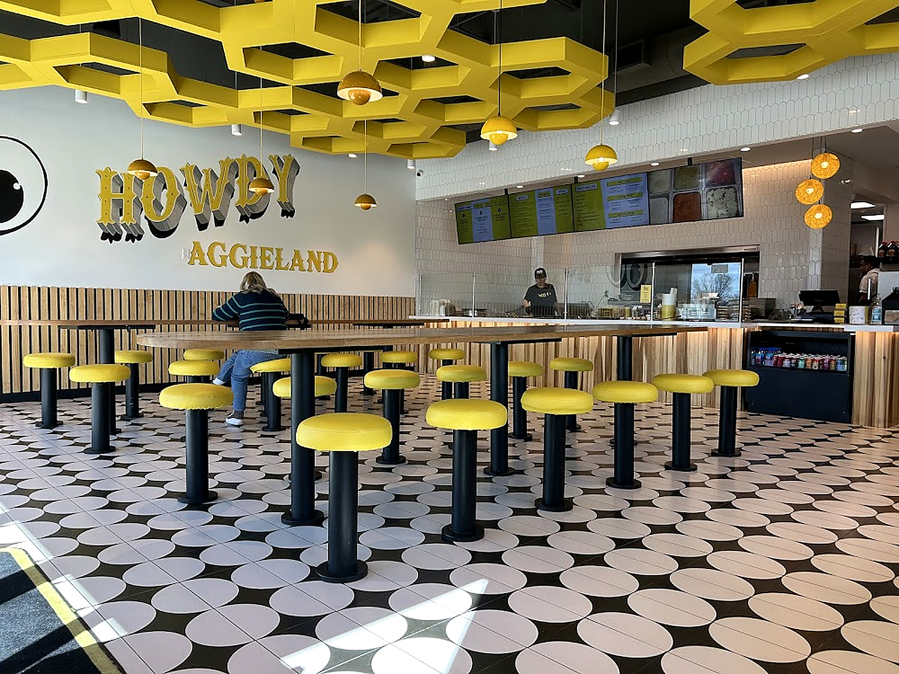 The dining room — long light-wood tables on black-and-white geometric tile, fixed stools with yellow cushions, and a sculptural yellow honeycomb ceiling feature with pendant dome lights