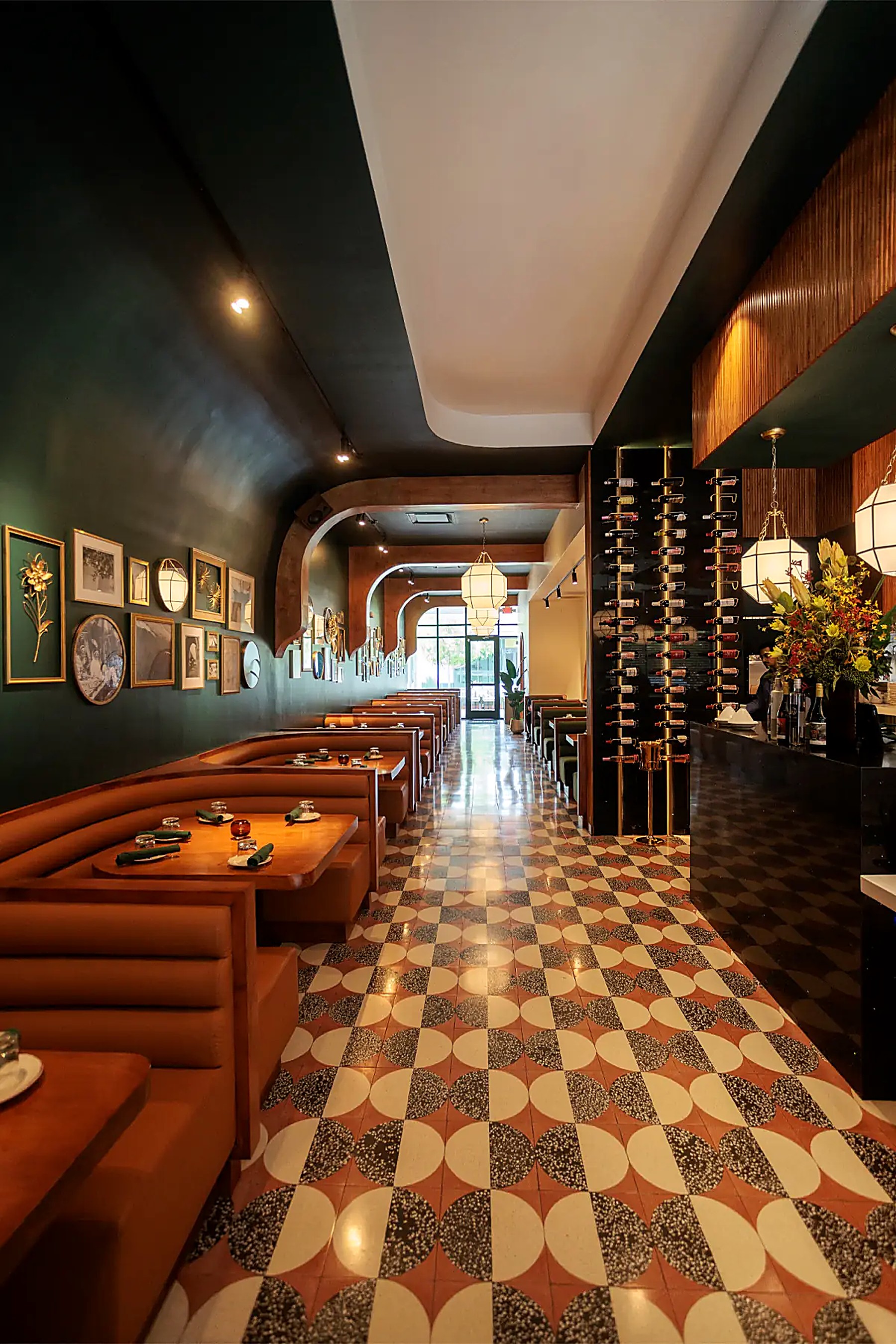 Portrait view down the dining aisle — brown leather booths along a dark green wall hung with framed pictures, patterned tile floor, and the bar with a tall wine display to the right