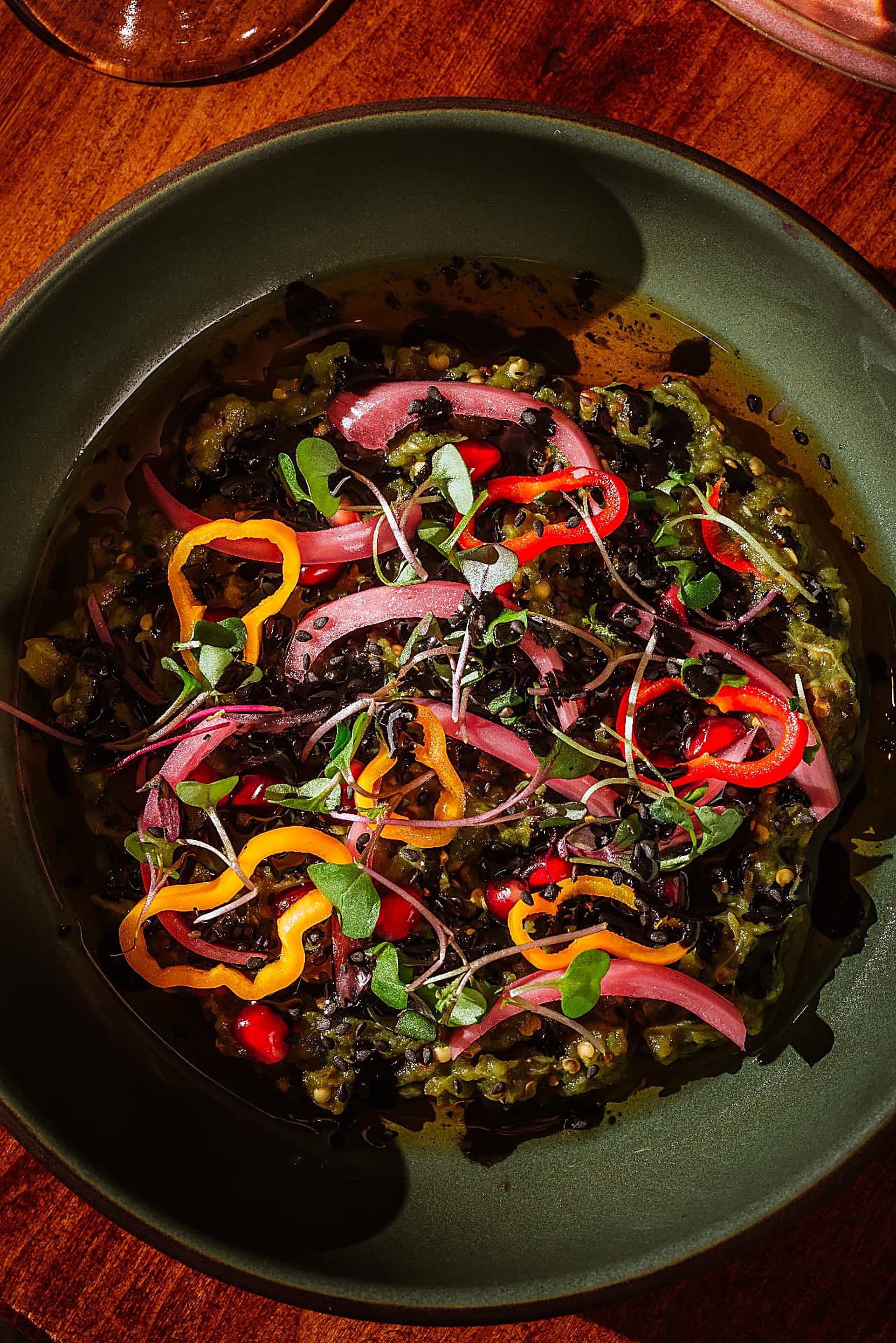A matte green bowl of herbed dip pooled with oil, topped with pepper rings, pickled red onion, pomegranate seeds and microgreens, on a warm wood table