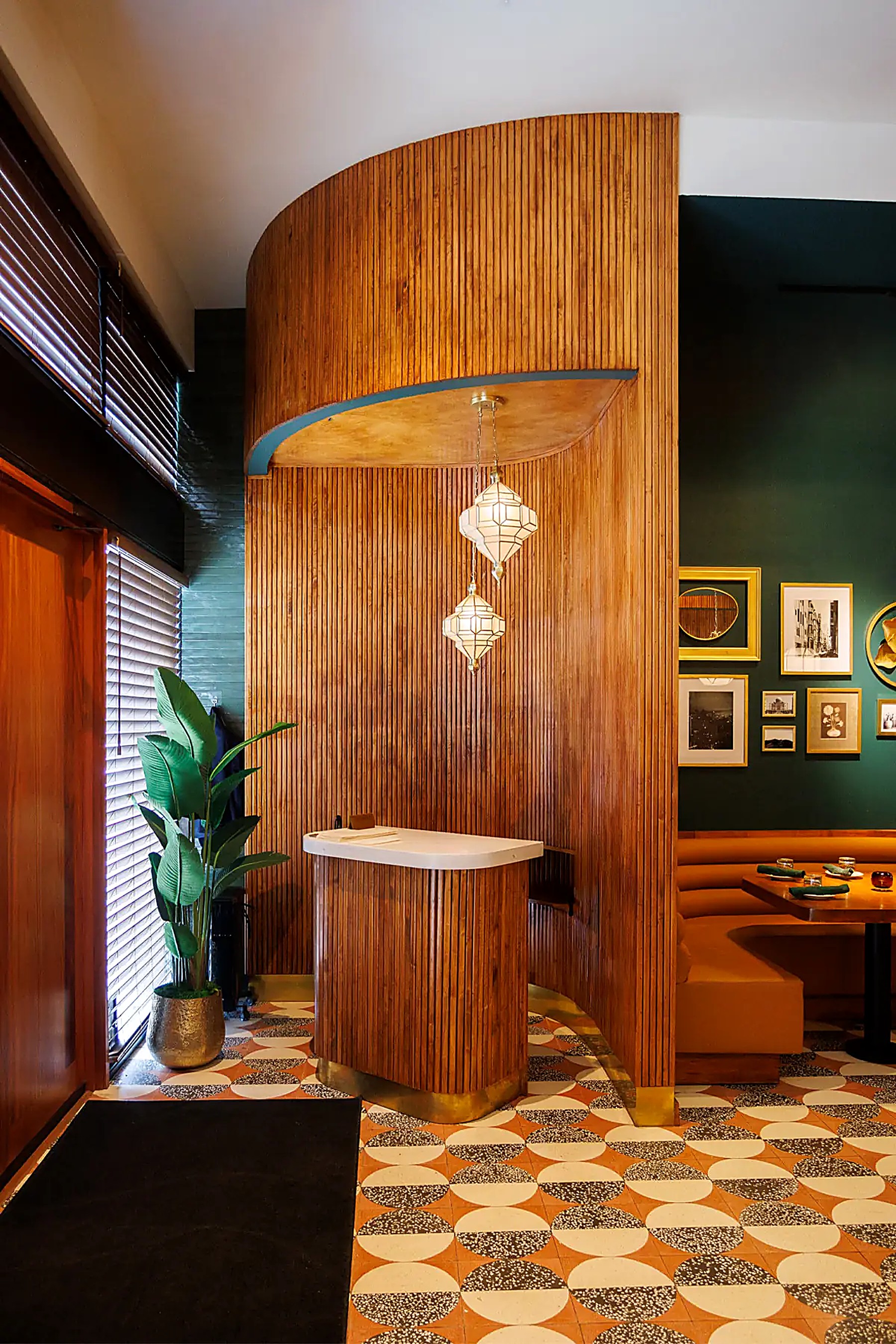 The entrance host stand — a curved wall of vertical wood slats beneath two geometric lantern pendants, beside a potted plant and the edge of a dining booth